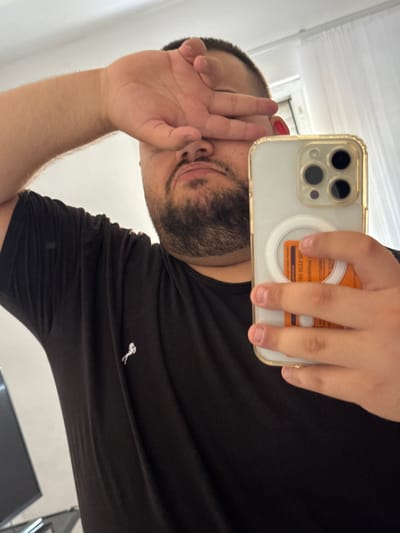 This is a selfie taken in a mirror, showing a person holding a smartphone. The person, who appears to be male, has dark hair and a beard, and is wearing a black t-shirt with a small white logo on the chest. He is covering part of his face with his hand, and his expression is difficult to discern due to the pose. The smartphone has a gold-colored case and a camera module with three lenses. A circular white accessory, possibly a phone grip or charger, is attached to the back of the phone, along with an orange card with some text. The background suggests an indoor setting with a white wall and a window with blinds. The lighting appears to be natural, indicating daytime. The orientation of the photo suggests it was taken in Byala Reka, Bulgaria, but there are no explicit location cues within the image itself.