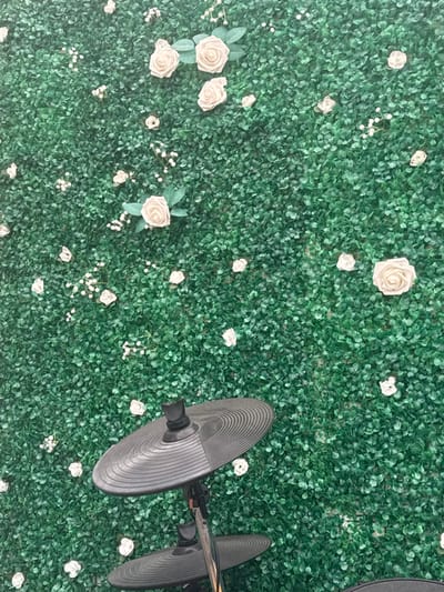The scene, reportedly in Neuville-aux-Bois, France, displays a vertical surface covered densely with artificial green foliage. Artificial cream-colored roses and clusters of small white artificial flowers are scattered across this green background. In the lower center, the upper section of an electronic drum kit's hi-hat stand is visible, featuring two black, ribbed cymbals mounted on a metallic support, with a black clutch mechanism at the top. No individuals are present in the image.