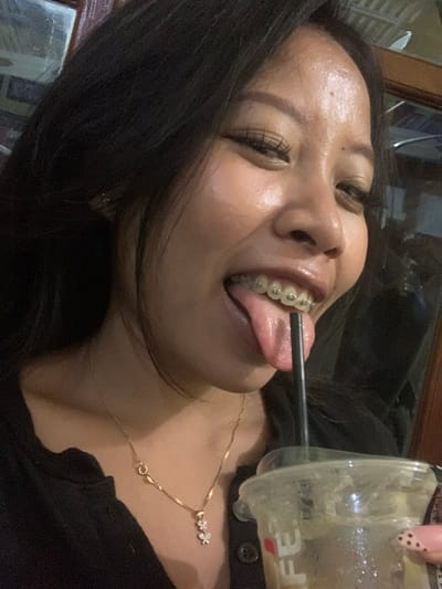 A medium shot captures a young woman with dark hair and braces, sticking her tongue out with a straw in her mouth, from a clear plastic cup. She wears a black shirt and a gold necklace with a small pendant. The background is blurred, but appears to be an indoor setting, possibly a cafe, with wooden elements. The lighting is dim, suggesting it might be evening. There are no visible indicators of the specific location within Jajag, Indonesia.