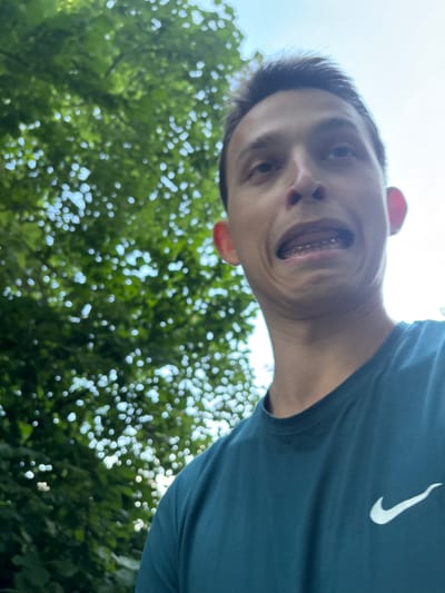 In this selfie, a young person with a wide, playful grin is captured from a low angle, looking up at the camera. Their mouth is open, revealing teeth and orthodontic braces, adding a touch of youthful individuality to the image. The person is wearing a dark teal t-shirt with a white Nike logo visible on the right side. The background is a blur of vibrant green foliage and bright white sky, suggesting an outdoor setting on a sunny day. The greenery, possibly from trees, is out of focus, creating a bokeh effect that draws attention to the subject's face. The lighting indicates it is daytime, likely late morning or early afternoon, given the brightness of the sky. The overall mood is lighthearted and energetic, conveyed through the person's exaggerated facial expression. The scene is set outdoors, possibly in a park or garden, with the sun filtering through the leaves, creating dappled light. There are no visible text elements other than the Nike logo on the shirt.