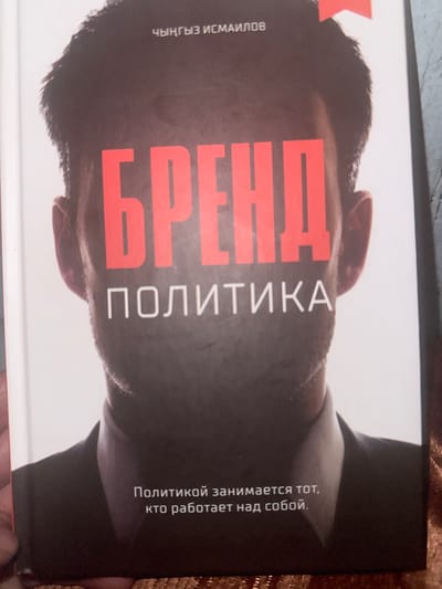 This is a close-up, top-down angled photograph of a book cover. The cover features a man's face, partially obscured by bold red text. At the very top of the cover, in white text, reads "ЧЫНГЫЗ ИСМАИЛОВ" which translates to "CHINGYZ ISMAILOV". Below this, also in bold red text, is the word "БРЕНД" (BRAND). Centered beneath that, in white text, reads "ПОЛИТИКА" (POLITICS). Towards the bottom of the cover, in smaller white text, is a quote that reads "Политикой занимается тот, кто работает над собой." (Only he who works on himself is engaged in politics.) The background is a plain white surface. There is no indication of the city of Sadovoe, Kyrgyzstan, in the image or its metadata.