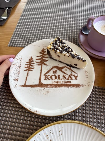 A slice of cheesecake is on a white plate with a chocolate powder design of trees and a house, along with the text "KÖLSAY LAKES TOWN". The plate is on a woven placemat. To the right, a lavender mug with a frothy drink sits on a matching saucer with a spoon. A fork is partially visible in the upper left corner. The scene appears to be in a cafe or restaurant. There is no discernible information about the location in the city of Kurmeti, Kazakhstan.