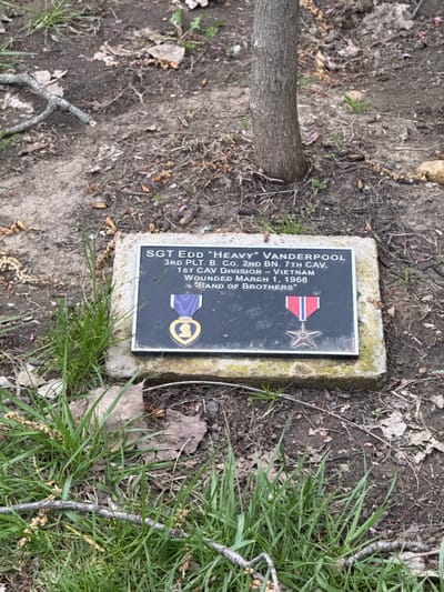 The image shows a grave marker for SGT Edd "Heavy" Vanderpool, a soldier who served in the 3rd Platoon, B Company, 2nd Battalion, 7th Cavalry, 1st Cavalry Division in Vietnam. He was wounded on March 1, 1968. The marker is placed on a concrete base and surrounded by dirt, grass, leaves, and small twigs. A small tree trunk is visible to the left of the marker. The location appears to be in Grove City, United States of America.