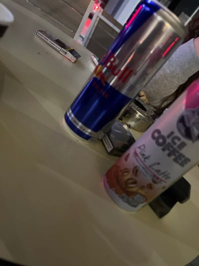 The image displays a close-up, angled view of a light-colored table surface in an indoor setting. On the table, a silver and blue Red Bull energy drink can is positioned prominently, leaning slightly to the right. To its right, a white and pink "Ice Coffee Pink Latte" can is visible in the foreground, also leaning slightly. Between these two cans, a metallic, circular ashtray containing dark residue is present. To the left of the Red Bull can, a white and red cigarette pack, bearing Bulgarian text, rests on the table. A second, darker cigarette pack is partially visible behind the "Ice Coffee" can, near the ashtray. A segment of a person's upper arm or shoulder, clad in a light grey long-sleeved garment, is visible on the far right edge of the frame. The background is out of focus, revealing indistinct indoor architectural elements and a red light source. The precise location within Piperkov Chiflik, Bulgaria, is not discernible from the visual evidence provided.