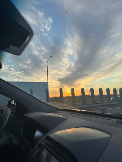 The image captures a view from inside a car driving towards a sunset, likely on the outskirts of Kushchovskaya, Russia. The sky is filled with dynamic clouds, ranging from wispy cirrus to heavier, grayer formations, with the sun peeking through in the distance, casting a warm, orange glow. The car's dashboard and steering wheel are visible in the foreground, with a hand on the wheel indicating the driver. Outside the car, a modern, white building is seen to the left, possibly a warehouse or industrial structure. A tall street lamp stands prominently against the sky. To the right, a series of dark, vertical bollards line a road or barrier, with faint silhouettes of distant buildings and potentially other vehicles. The overall impression is one of travel and the beauty of a rural or semi-urban sunset.