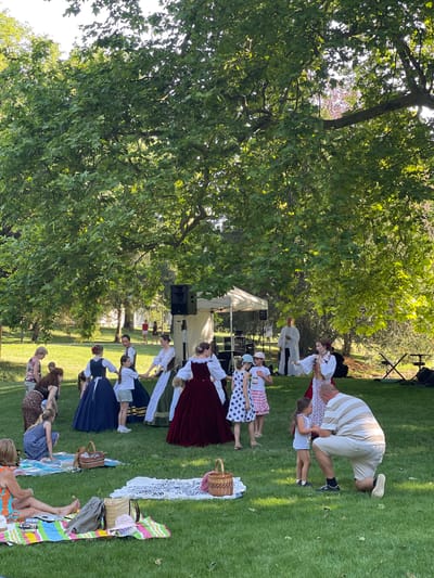 This media file depicts an outdoor performance or gathering in a grassy park with large trees. The sun is shining, suggesting it is daytime. Several people are dressed in what appear to be historical or traditional costumes, with women in long dresses and head coverings, and men in suspenders and caps. Some of the women are holding hands, seemingly participating in a dance or a structured activity. There are also several children present, some of whom are also dressed in similar attire.
In the background, there is a white tent structure and some audio equipment, indicating a performance or event. Spectators are seated on blankets spread out on the grass, some with picnic baskets. The overall atmosphere appears festive and relaxed. The presence of trees, sunlight, and open green space suggests a pleasant outdoor setting. The individuals in costume are the primary subjects, with the focus on their interaction and the event they are part of. The scene is set in Stupava, Slovakia.