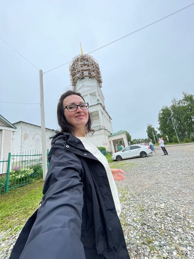 In Кстинино, Russia, a woman with dark hair and glasses takes a selfie outside a white building that appears to be a church under renovation. The building has a tall, multi-tiered structure, with the upper section covered in scaffolding, topped with a golden spire. To the left of the church is a smaller building with pale pink siding and a white trim. A green metal fence separates a grassy area with some flowering plants from the main path. The ground is covered in gravel. In the background, a white car is parked, and several people are visible, including a man holding a child. Tall green trees line the far side of the gravel area. The sky is overcast and grey.