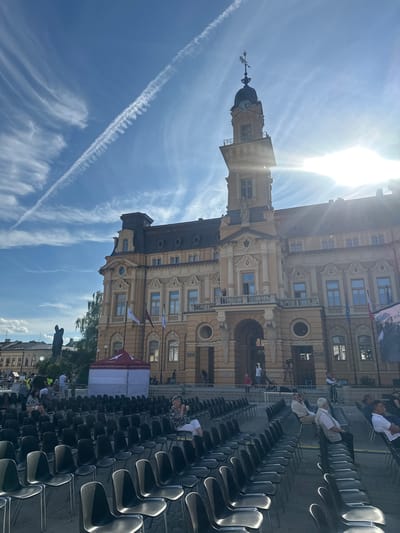The image shows an outdoor event setup in front of the Sokół building in Nowy Sącz, Poland. Rows of empty black chairs fill the foreground, with some people seated in them in the middle ground. The Sokół building, an ornate yellow structure with a clock tower, dominates the background. The sky is blue with wispy clouds and a prominent contrail. A red and white tent is set up in front of the building, and a statue of a figure is visible to the left of the building. The sun is shining brightly, creating lens flare in the upper right portion of the image.