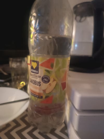A plastic bottle of Edeka "Herzstücke Jakobus" apple juice is the primary subject, positioned centrally in the frame. The label features a yellow heart icon, the Edeka logo, and imagery of apples with geometric patterns. The bottle contains a reddish-brown liquid, suggesting it is juice. In the background, out of focus, are what appear to be kitchen appliances or containers, including a white object with defined edges on the right and a dark, possibly black, appliance behind it. To the left of the bottle, a white plate with a dark rim is visible, resting on a patterned surface resembling a tablecloth or placemat with a chevron design. The overall lighting suggests an indoor environment, possibly a kitchen or dining area, with reflections on the plastic bottle indicating ambient light sources. The specific location in Freiburg im Breisgau, Germany, cannot be determined from the visual information provided.
