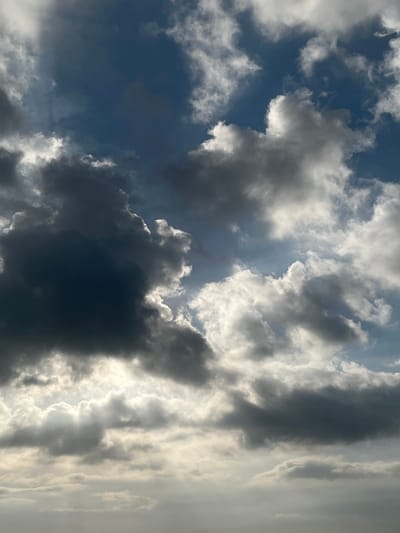 The image is a low-angle shot of a cloudy sky with the sun peeking through. The clouds range in color from dark grey to bright white, creating a dramatic contrast. Some clouds appear wispy and translucent, while others are dense and voluminous. The sunlight filters through in rays, illuminating portions of the clouds and casting a soft glow on the sky. There are no people, subjects, objects, or actions depicted in this image; it is solely a view of the sky and clouds. The scene does not provide any specific indicators of the city of Tambakreja, Indonesia.