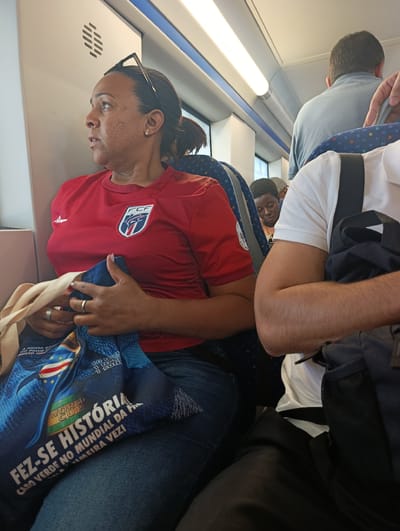 In Seixal, Portugal, on a train, a woman with dark hair pulled back is seated on the left, looking out a window. She wears a red t-shirt with a crest and is holding a blue bag with white text that reads "FEZ-SE HISTÓRIA CABO VERDE NO MUNDIAL DA P." A pair of glasses rests on her head. To her right, a man in a light blue shirt faces away from the camera. A man in a white polo shirt sits to the right, with a black backpack visible. The train car interior features beige walls and blue seats, with overhead lighting illuminating the scene.