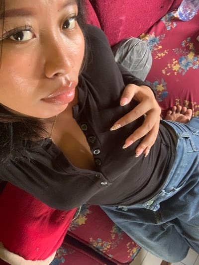 The image shows a close-up, overhead view of a person lying down on a red floral patterned sofa. The person has dark hair and is wearing a black long-sleeved shirt with buttons down the front and blue jeans. Their left hand is resting on their chest, displaying nails painted with a light pink base and small brown dots. The person's face is partially visible, with their eyes looking towards the camera. Their lips are glossy and appear to be slightly parted. To the upper right of the image, a partially visible clear plastic bottle with a blue cap is on the sofa. The overall setting is an indoor environment, consistent with a living room or lounge area. The image does not provide any specific information or visual cues to determine a location within the city of Jajag, Indonesia.