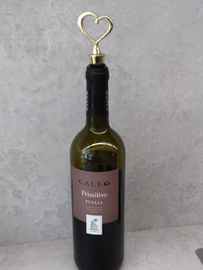 A bottle of Cale Primitivo Puglia red wine lies on a light gray, mottled surface, possibly a countertop or table. The bottle is dark green glass and is uncorked, with a gold-colored wine stopper featuring a heart-shaped handle inserted into the neck. The stopper is the most prominent object in the foreground. The wine label is a dusty rose color and has text printed vertically and horizontally. Visible text includes "CALE" at the top, followed by "Primitivo" and "PUGLIA" below it. A smaller white label with some text and a crest is also visible on the right side of the main label. The background is a textured, light gray surface that appears to be a stone or marble material. The lighting is even, suggesting indoor lighting. No people are present, and the scene is static, with no activity taking place. The overall impression is that of a product shot or a still life.