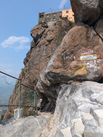 The image displays a steep, rocky landscape located in Kadaladi, India, under a clear blue sky with minimal cloud cover. The scene is dominated by large, weathered boulders exhibiting shades of brown, grey, and reddish-brown, with visible cracks and some white mineral streaking. Numerous rocks in the mid-ground are covered with white, red, and yellow graffiti, including discernible names such as "B BALU," "V HARISH," "ANBU," and "M_NOVOTH." A small red marking is affixed to a rock on the far right.

A multi-story structure, appearing to be two to three stories high with dark grey lower sections and a lighter, possibly orange, upper section, is situated precariously atop the highest rocky peak in the background. The building's roofline features a visible defensive barrier, possibly barbed wire.

In the foreground and mid-ground, a narrow, unpaved pathway, seemingly carved directly into the rock, winds towards a dark, tunnel-like passage between large boulders on the right side of the frame. To the left of this path, a metal mesh fence, showing signs of damage including a large tear, is erected on concrete bases with metal posts, presumably guarding a drop-off to a lower elevation or valley visible in the distant background. Rough-hewn steps are present in the immediate rock foreground on the right. Sparse, dry vegetation grows in crevices between the rocks. No identifiable individuals are clearly visible within the scene.