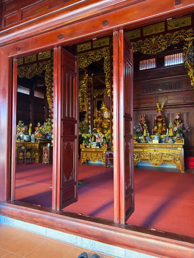 The image depicts the interior of a temple in Diên Lạc, Vietnam. The viewpoint is from an entryway framed by wooden beams and open doors, revealing a red carpeted room with ornate golden altars and tables laden with offerings of flowers, fruit, and objects. Several statues are placed on the altars. The interior is richly decorated with carved wooden details. The light appears to be natural, illuminating the interior of the temple.
