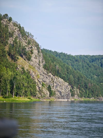A river flows in the foreground of the image. The water is calm with a slight ripple. On the left side of the image, a steep, rocky cliff rises from the water's edge. The cliff is covered in lush green trees, primarily pines and birches, with patches of grass and smaller vegetation at its base. The cliff face itself is a mixture of grey rock and darker soil, with some trees growing precariously on ledges.
To the right of the cliff, the landscape continues with a densely forested hillside that slopes upwards and recedes into the distance. This hill is also covered in a thick carpet of green coniferous trees, creating a textured, undulating green mass. The sky above is a pale, clear blue with a few faint, wispy clouds and some tiny specks that could be birds or insects.
The scene depicts a natural landscape, likely a river valley bordered by steep, forested cliffs. The light suggests it is daytime, possibly late morning or early afternoon, with soft sunlight illuminating the scene. There are no people or man-made structures visible in the image. The overall impression is one of a serene, untouched natural environment.