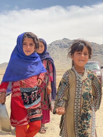 Three young girls stand outdoors in Ashoqah, Afghanistan, in front of a desert landscape with a mountain in the background. The girl in the foreground on the right wears a patterned dress with a beaded necklace. The girl on the left wears a red outfit with a blue headscarf. She also carries a metal object. The girl in the middle wears a floral dress and a blue headscarf. A red vehicle is visible in the background.