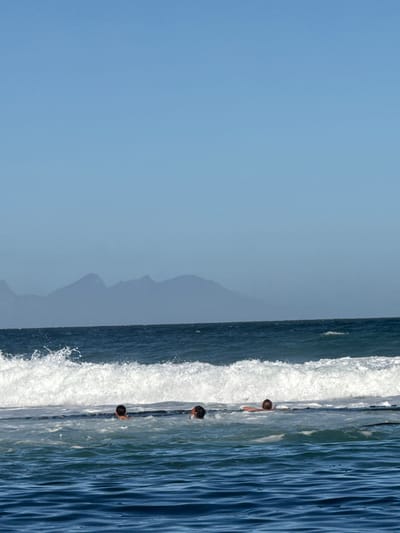 The image shows three people in the water at Muizenberg beach, South Africa. A wave crashes beside them. In the background, mountains are silhouetted against a pale blue sky. The image has been rotated.