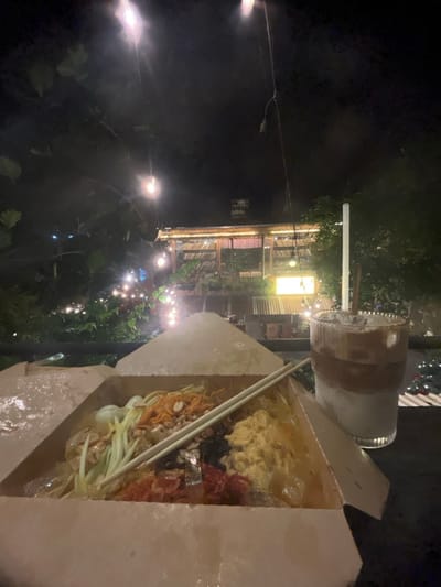 The image depicts a nighttime outdoor dining scene in Cần Thơ, Vietnam. In the foreground, a light-colored, open cardboard takeout box contains a Vietnamese dish, likely a salad or noodle preparation, featuring shredded green components, peanuts, dried red strands, and other mixed ingredients. A pair of white chopsticks rests diagonally across the dish. To the right, a ribbed clear glass holds a layered iced coffee drink, showing distinct white (milk) and brown (coffee) sections with ice, and a clear straw. These items rest on a dark, reflective surface. The background showcases a multi-tiered wooden structure with slatted roofs and open sides, illuminated by numerous warm-toned string lights. Lush green foliage and trees are interspersed throughout the scene. A bright yellow sign is visible on the lower level of the background structure, above a corrugated metal roof. The sky is dark, indicating night, with faint distant lights visible. No individuals are present in the frame.
