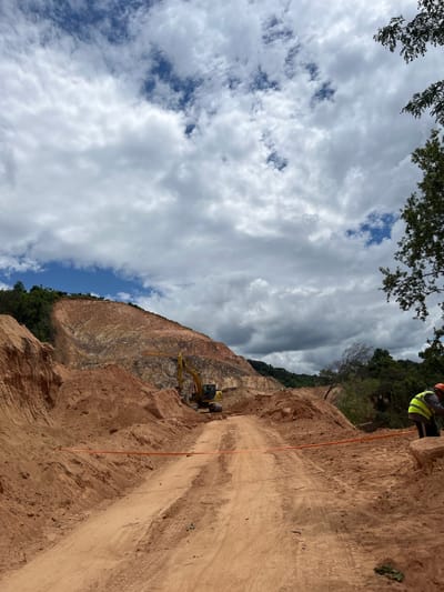 A yellow excavator is visible in the middle of a dirt road leading up a steep, red-soiled mountainside. The mountainside shows layers of exposed rock and dirt, indicating excavation or mining activity. Two construction workers, wearing high-visibility vests and hard hats, are present on the right side of the road, working near a large boulder. An orange safety ribbon is stretched across the road in the foreground. The sky is filled with dramatic, grey and white clouds against a backdrop of blue sky. Lush green trees are visible on the left and right edges of the frame, with more trees and foliage further up the mountainside. The overall scene depicts an active construction or earthmoving site, possibly a quarry, located in a hilly, natural environment, with elements of construction work in progress under a cloudy sky. The description does not specify the city of Son Dien, Vietnam, as this information is not visually derivable from the image.