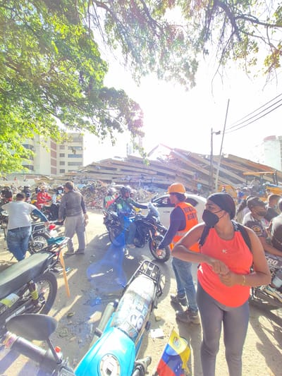 In Caraballeda, Venezuela, a group of people are gathered near a pile of rubble that appears to be the remains of a collapsed building. Some individuals are wearing safety vests and hard hats, suggesting they are involved in rescue or recovery efforts. Several motorcycles are parked in the area, and a few people are seated on them. A yellow excavator is visible in the background near the debris. The scene is lit by bright sunlight filtering through trees. A Venezuelan flag is propped up near the foreground.
