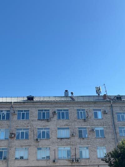 The image shows the facade of a multi-story brick building under a clear blue sky. The building has multiple rows of windows, many of which appear to be double-paned. Several air conditioning units are mounted on the exterior walls. The roof of the building is visible, featuring a sloped, metallic surface with visible seams. On the roof, there is a small dormer window and a telecommunications antenna structure. The scene is devoid of people and any discernable activity. This building is located in the city of Belovo, Russia, and it appears to be a commercial or administrative building.