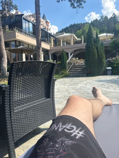 The image depicts a sunny day at a resort in Ognyanovo, Bulgaria. In the foreground, a person is relaxing on a lounge chair, wearing black shorts with a graphic print. Their legs and feet are visible, with a tattoo on one ankle. To the left, a black wicker chair is partially in frame. The background features a multi-story building with a castle-like architectural element and large windows, surrounded by trees and a paved area. Several outdoor umbrellas and seating areas are visible on a terrace. The environment suggests a leisure setting, possibly a hotel or spa, with a mountain or hillside backdrop.