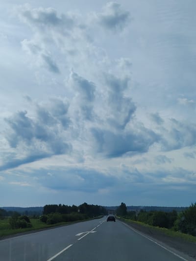 The image depicts a road in Shavkunovo, Russia. The road is asphalt, with a white directional arrow painted on the left lane. A car is driving on the road. The background is a landscape of trees and fields. The sky is cloudy, with gray and white clouds.