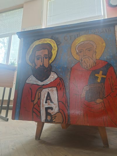 A vibrant, hand-painted wooden icon stands upright on a simple wooden easel or stand, positioned indoors in what appears to be a well-lit room, possibly a classroom or community hall. Large windows with horizontal blinds are visible to the left, letting in bright natural light, while a wooden parquet floor is seen below.
The icon itself depicts two male figures, consistent with Orthodox Christian iconography. Both saints are depicted with golden halos around their heads and possess calm, serene expressions.
The figure on the left has a white beard and mustache, fair skin, and wears a red robe adorned with a prominent yellow cross on his left shoulder. He holds a dark, closed book in his hands. Above him, faintly inscribed in Cyrillic, are the letters "СТ. И МЕТОДИЙ", identifying him as Saint Methodius.
The figure on the right has a dark beard and hair, fair skin, and wears a red robe over a white undergarment. He holds an open white book or scroll, on which clearly visible Cyrillic letters spell "АЗЪ" (Azъ), the Old Church Slavonic word for "I" and the first letter of the Glagolitic and Cyrillic alphabets. This inscription, combined with the presence of St. Methodius, strongly identifies him as Saint Cyril, the co-creator of the Slavic alphabet.
The background of the icon is a mottled blue, creating a spiritual atmosphere. The artistic style is somewhat naive or folk-like, characterized by bold outlines and simple coloring.
Faint Cyrillic text, "...ИЛИАДА" (Iliada), is also visible on a white surface near the window to the right of the painting. Given the location context of Dolni Chiflik, Bulgaria, the depiction of Saints Cyril and Methodius, the revered Enlighteners of the Slavs, is culturally significant.