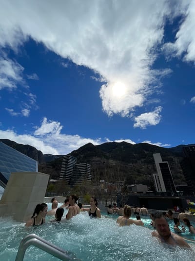 The image shows a group of people in a pool. The pool appears to be located outdoors, with mountains and buildings in the background, including several modern high-rise buildings. A man faces the camera at the edge of the pool. The sky is blue with scattered white clouds and the sun. The building visible is the Caldea, Thermoludic Center, Les Escaldes, Andorra.