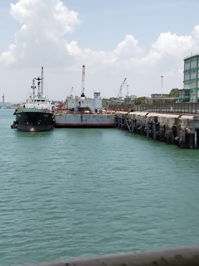 A port scene in Mkomani ward, Kenya, is depicted under a partly cloudy sky. The foreground features a blurry grey-white railing above a body of teal-green water displaying small ripples. In the midground, a dark green-hulled vessel, identified as "TAURUS," is moored. Directly adjacent to it and against a concrete pier is a grey flat-decked barge, partially obscured, with the words "FUEL DANGER" visible on its side. Multiple industrial cranes, some with red tips, are visible on and behind the barge and pier, indicating active operations. The robust concrete pier extends to the right, fitted with blue vertical and black horizontal fendering elements. Small, indistinct figures are visible on the barge and pier. In the background, a multi-story building with green and white sections stands on the far right, with other industrial structures and a distant bridge-like formation visible on the horizon to the left.