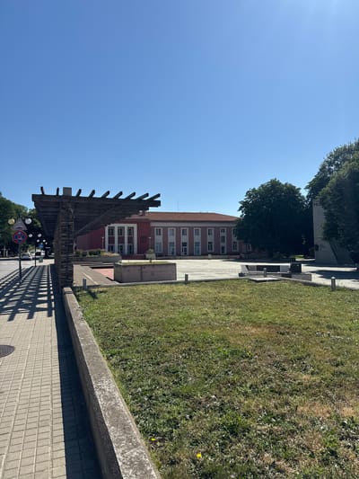 The image depicts an outdoor scene in the city of Shabla, Bulgaria, on a clear, sunny day. In the foreground, a paved walkway with a checkered pattern is visible on the left, casting a geometric shadow from a wooden pergola structure. A concrete border separates the walkway from a grassy area to the right. The pergola itself is constructed with weathered wooden slats supported by stone pillars.
In the middle ground, a red building with a tiled roof and numerous windows dominates the scene. The building has a neoclassical architectural style with prominent white columns supporting an overhang. In front of the building, there are some low-lying bushes and a paved area. To the right of the red building, a large, dark green tree stands tall, and further to the right, a tall, light-colored monument or sculpture is partially visible.
The background is a clear, bright blue sky. Traffic signs are visible on the left side of the walkway, including a traffic light and a circular no-entry sign. A white car is also seen on the road to the left. The overall environment suggests a public space, possibly a town square or a park area adjacent to a civic building.