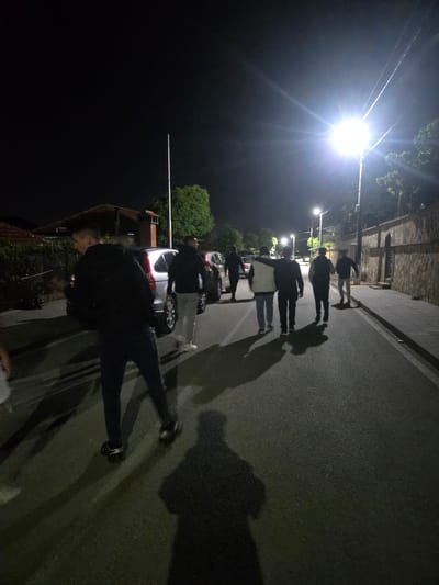At night, a group of individuals walks along a paved road in what is potentially Pripek, Bulgaria. Streetlights cast long shadows from the people and parked vehicles along the road. The surroundings are dark, with glimpses of buildings and trees. The people are moving in the same direction.