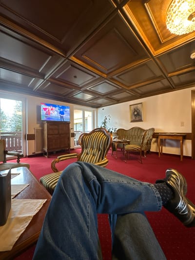 Inside a room featuring wood-paneled walls and a red carpet, a person wearing jeans and black shoes with white soles rests their legs. A television screen displays a colorful scene. Striped chairs are arranged in the room, and windows offer a glimpse of the outside. A table with a light-colored cloth is partially visible in the foreground. A chandelier hangs from the ceiling, providing illumination.