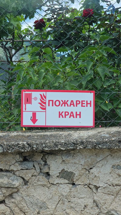 A rectangular sign with a red border and a white background is attached to a chain-link fence. The sign features a graphic of a fire hose and a flame symbol on the left side. To the right of the graphic are two lines of text in Cyrillic script. The top line reads "ПОЖАРЕН" in large, red capital letters, and the bottom line reads "КРАН" in smaller, red capital letters. The sign appears to be a warning or informational sign indicating a fire hydrant. Behind the sign, lush green foliage and the branches of a rose bush with red roses are visible, with a glimpse of the sky and a distant hillside. The sign is mounted on a stone wall. The overall setting suggests an outdoor environment, possibly a garden or park area.