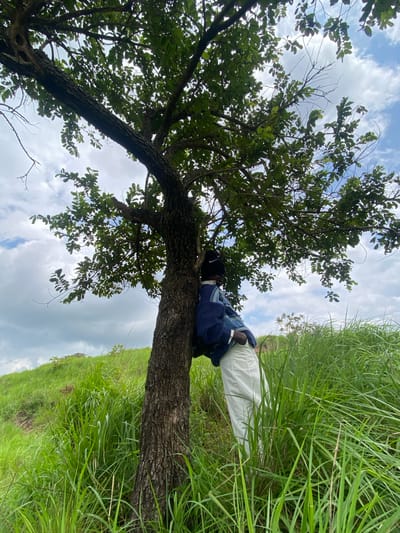 A person leans against a tree trunk, embracing it with their arms. They are wearing a dark beanie, a blue knitted sweater with a patterned design, and white pants. The person is positioned on a grassy slope with tall, green grass surrounding them. Above, the branches of the tree are covered in leaves, and the sky is visible with scattered white clouds. The scene appears to be outdoors, in a rural or natural setting, potentially in Ebuwana, Nigeria. The overall impression is one of solitude and connection with nature.