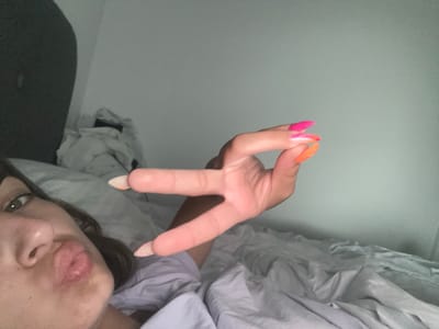 A young woman is lying in bed, looking at the camera and making a peace sign with her hand. She has long, pointed fingernails painted in bright pink and orange. The room appears to be dimly lit, with a white wall behind her. The overall impression is one of a casual, personal moment captured on camera. The location is not specified in the image.