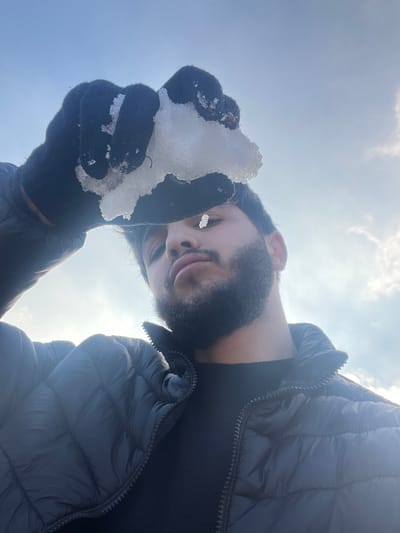 A male individual is depicted outdoors in Talfita, Syria, against a bright, cloudy sky. The subject, a young adult male with dark hair, a beard, and a mustache, is wearing a black puffer jacket over a black long-sleeved shirt and black gloves. He holds an irregularly shaped piece of white, semi-translucent snow or ice in his gloved left hand directly above his head, partially obscuring the upper portion of his face, including his forehead and eyebrows, from the camera's perspective. A smaller fragment of snow or ice is visible suspended directly in front of his face, roughly at eye level. The subject is looking directly towards the camera with a neutral expression. The background consists of a clear, light blue sky with scattered white clouds, illuminated by daylight.