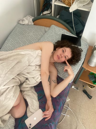 A person with curly hair is lying in bed, partially covered by a white blanket. They are holding a white smartphone in their right hand and pointing to their right eye with their left index finger. A black tablet is situated on the pillow next to their head. The bed has a patterned gray and white pillowcase and a blue-purple patterned sheet. Next to the bed is a white drawer unit containing various clothing items. A small side table with a metal cylinder, green containers, and various items is visible above the bed. The floor is tiled and strewn with wires, a tube of ointment, and other small objects.

The image does not provide sufficient information to verify the location in Le Pré-Saint-Gervais, France.