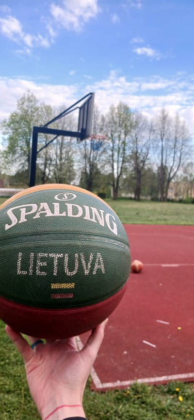 The image shows a Spalding basketball with the word "LIETUVA" (Lithuania) and a depiction of the Lithuanian flag. The basketball is held by a hand with a red wristband. Behind the basketball, there is a red basketball court with a basketball hoop, trees, and a cloudy sky. The location is likely Butkiškė, Lithuania, based on the inscription on the basketball.