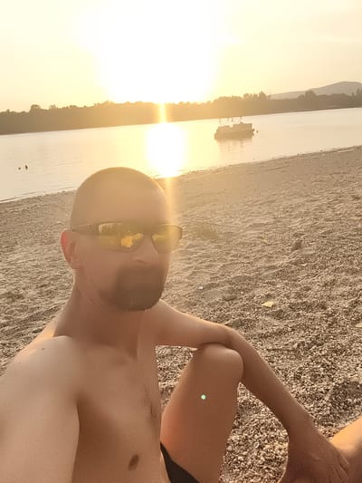 This is a selfie of a man sitting on a pebble beach with a lake and sunset in the background. The man is shirtless and wearing sunglasses, and his face is partially obscured by the glare from the sun. He has a beard and short hair. In the background, a pontoon boat is visible on the water, and a treeline stretches across the horizon. The sun is setting, casting a warm, golden glow over the scene. The sky is bright with the sun's rays, and the water reflects the light. The beach appears to be made of small pebbles and sand. The overall mood of the image is peaceful and relaxed, suggesting a pleasant end to a day outdoors. The location is identified as Dunakeszi, Hungary.