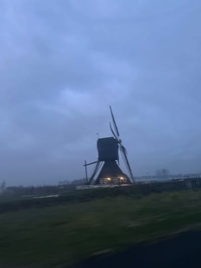 The image shows a windmill at dusk in Gellicum, Netherlands. The windmill is the dominant object, featuring a dark, box-like structure on a raised base with the characteristic blades extending outwards. The sky is overcast and gray, suggesting a gloomy atmosphere. To the right of the windmill, there is a field of green grass, and a body of water. The image is slightly out of focus and the windmill is angled relative to the frame, adding to the sense of movement or perspective.