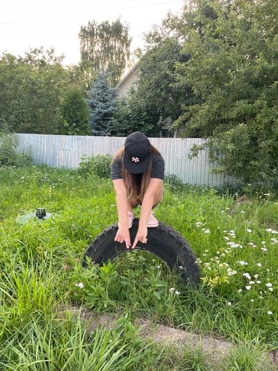 A young person wearing a black baseball cap and a dark t-shirt is crouched down next to a large, worn-out tire in a grassy area. They are wearing light pink sneakers. The person is reaching out with their right hand towards the tire, with their fingers extended. The scene is outdoors, with lush green grass and scattered white wildflowers. In the background, there is a metal fence and a dense canopy of green trees, with a building visible through the foliage. The lighting suggests it is daytime, possibly late afternoon or early evening, with a bright sky. The overall impression is one of a casual, outdoor activity in a rural or semi-rural setting, likely in Pushkino, Russia. There is no visible text in the image.