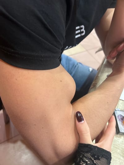 In Mirkovo, Bulgaria, an indoor scene captures an intimate moment involving two individuals. The primary focus is on the arm of one person, adorned in a black short-sleeved t-shirt, featuring a visible, flexed bicep. This arm exhibits light to medium skin tone with fine hairs and a few small moles or freckles. The second person's hand, covered by a black lace fingerless glove and featuring dark, possibly burgundy, polished fingernails, gently rests on the bicep of the first person.
The person wearing the t-shirt is seated, or at least positioned such that their torso, clad in the black fabric, is visible. A white, stylized logo on the t-shirt prominently displays what appears to be an uppercase "M" followed by two vertical bars (resembling "MII" or "MП"). In the background, hints of blue jeans with a torn detail suggest the lower body attire of the second individual. Further back, a light-colored surface, possibly a tiled floor or table, and a partially obscured object resembling a box or package are visible. Text on this object includes "SOBIYA" or "SOBIA". The overall setting appears casual and relaxed.