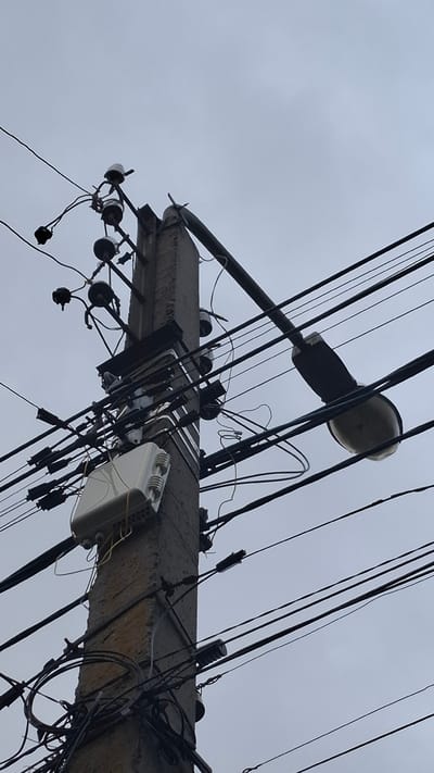 This image, taken in Rassvet, Russia, depicts a concrete utility pole set against an overcast, light grey sky. The pole is heavily laden with numerous overhead utility lines of varying thicknesses, predominantly dark in color. Towards the top, multiple white and dark ceramic or porcelain insulators are visible, securing several sets of electrical wires. Below these, a metallic bracket supports additional lines and components.
Mid-pole, a prominent white, rectangular enclosure, likely a telecommunications or fiber optic distribution box, is securely mounted. Several thin yellow wires emerge from the bottom of this box, intertwining with other dark cables that run vertically and horizontally along the pole. Further down, numerous black plastic strain relief clamps and cable bundles are attached, managing the intricate web of wires.
Extending horizontally from the right side of the pole is a dark-colored street light fixture, equipped with a large, oval-shaped lamp, illuminating the unseen area below. The pole itself appears weathered, exhibiting a rough, textured surface. No people, animals, or other distinct environmental features are visible in the frame, which focuses solely on the utility infrastructure.