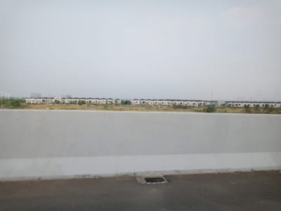 In Kosambi Barat, Indonesia, a clear sky hovers over a residential development and an overpass. The foreground is dominated by a concrete barrier and a grey asphalt road with a storm drain. Beyond the barrier, a row of identical houses with light-colored facades, dark arched roofs, and individual garages stretches across the horizon. A line of trees and green vegetation separates the houses from the road. In the distance, a few taller buildings rise, and a cell tower is visible. The overall scene suggests a modern suburban area under a bright, overcast sky.