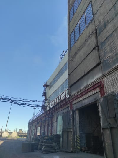 The image depicts an industrial building with a clear blue sky in the background. The building features a multi-story structure made of concrete and brick, with rows of windows on the upper levels. A metal fire escape is visible on one side of the building. In the foreground, there is an open loading bay with heavy industrial doors. Pallets of materials are stacked near the loading bay. Electrical cables are strung across the upper part of the scene. In the distance, industrial elements and what appear to be large circular structures are visible, along with a construction crane. The overall impression is one of an active industrial or manufacturing site. This scene is not in the city of Kyzyl, Kazakhstan; it is likely an industrial area elsewhere.