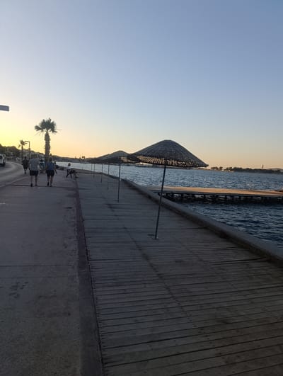 This media file shows a waterfront promenade at dusk in Urla, Turkey. Several people are walking along the paved walkway on the left, which borders a body of water. A couple in the foreground are holding hands and walking away from the camera. Further down the walkway, another person is seated on a bench.

To the right of the walkway is a wooden pier extending into the water. Several straw umbrella-like structures are positioned along the pier, suggesting a recreational area. The water is calm with gentle ripples. In the distance, across the water, a shoreline with buildings and trees is visible, with a tall structure that might be a lighthouse or a tower. The sky is clear with a gradient of pale blue to a warm yellow near the horizon, indicating the setting sun.

The overall scene evokes a peaceful and relaxed atmosphere, typical of an evening stroll by the sea. There is no visible text in the image.