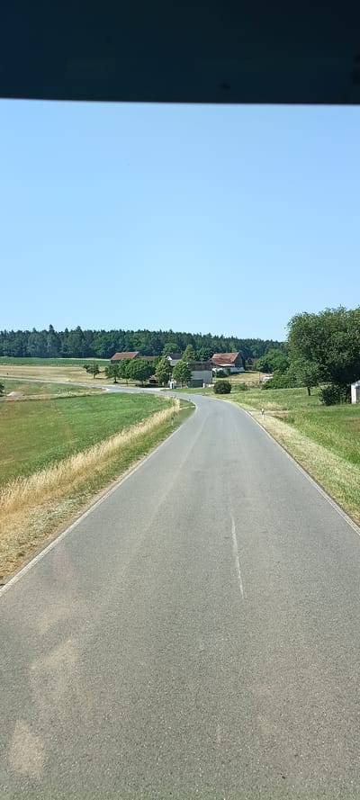 The image depicts a view from a vehicle on a paved road that curves to the right. The road is bordered by grassy fields with some dry, golden grass along the edges. In the distance, a cluster of buildings, likely part of a farm or rural settlement, is visible. These buildings have red-tiled roofs and are surrounded by trees. A dense line of trees forms the background, with a clear blue sky above. The scene appears to be a rural setting in the vicinity of Vilseck, Germany. There are no people or active events visible in the image.