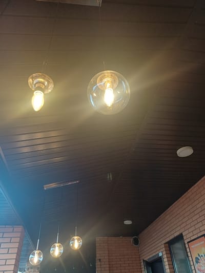 The image shows the interior of a cafe, likely in Ukrainka, Ukraine, with a focus on the ceiling and lighting. The ceiling is paneled with dark wood. Several pendant lights are visible, including a single, clear globe-shaped light with an Edison-style bulb, and a cluster of three similar lights hanging lower. Two of these globe lights are illuminated. There is also a single, smaller Edison-style bulb hanging without a globe. A circular white ceiling light fixture is present in the upper right portion of the image. The walls are constructed of light brown brick. A poster with text that appears to be in Russian or a Cyrillic-based language is visible on the far right. The scene is dimly lit, with the primary illumination coming from the pendant lights.